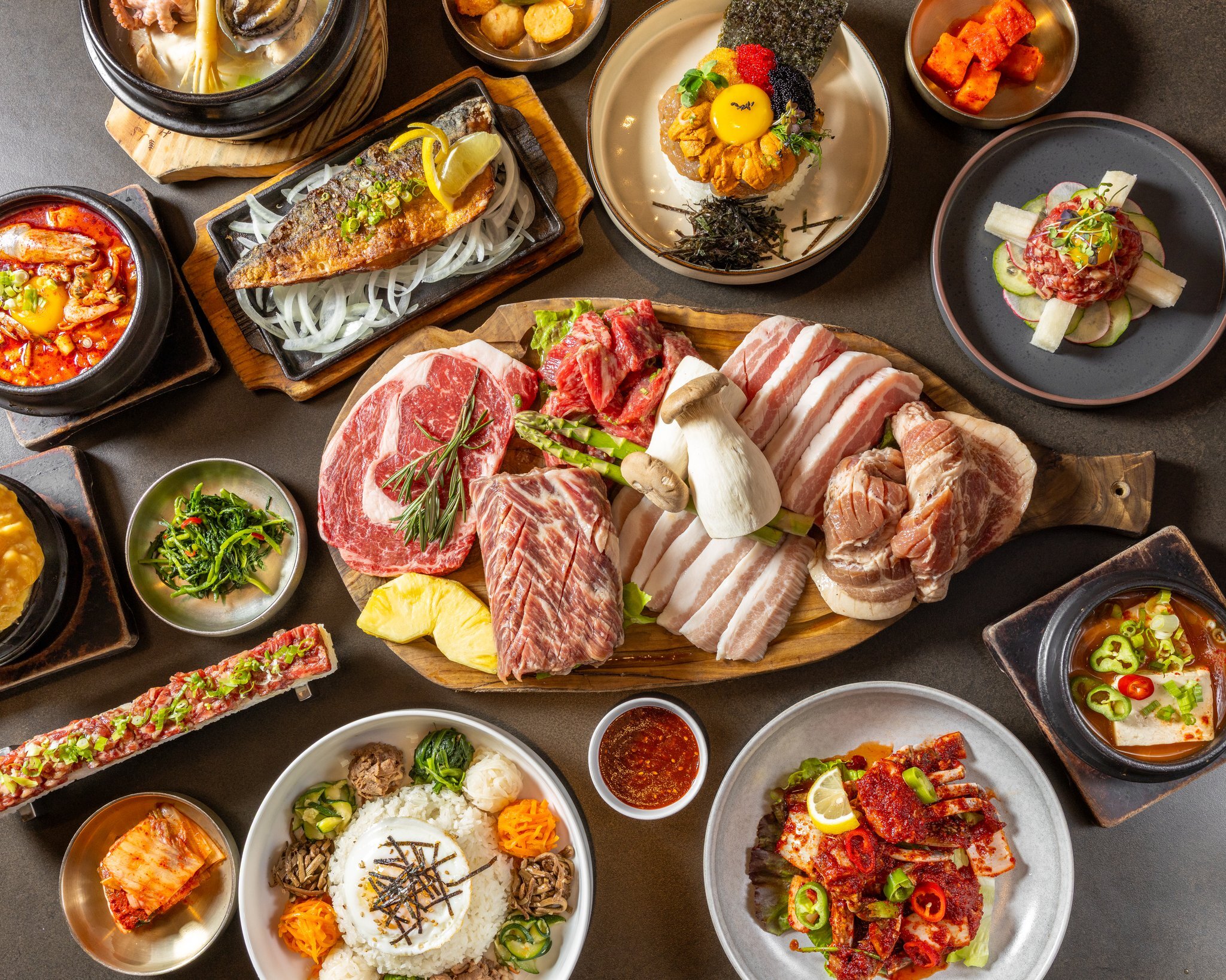 The table loaded end to end, the grill board at the center with stews and bibimbap all around